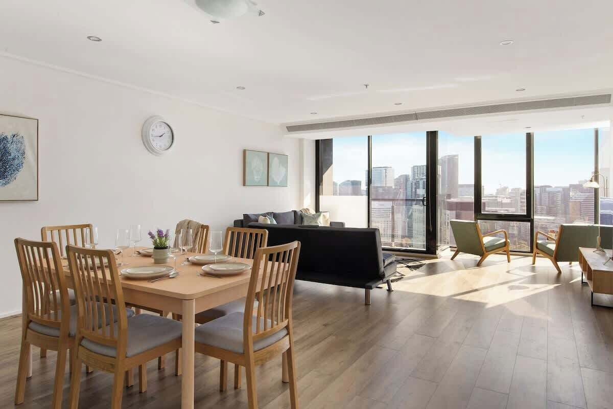 A spacious living area with a dining table, chairs, a sofa, and a large window offering a city view.