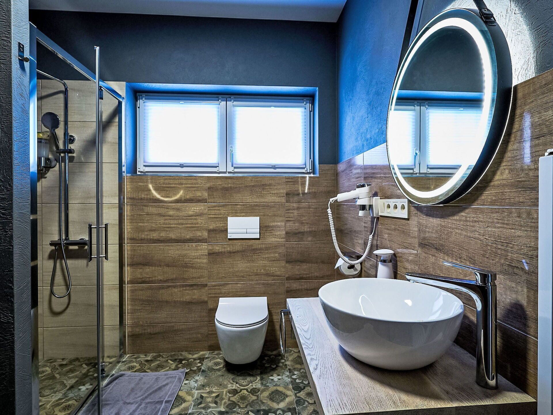 Une salle de bain moderne dotée d’une douche à l’italienne, de toilettes, d’un lavabo avec un grand miroir et d’une fenêtre laissant entrer la lumière naturelle.