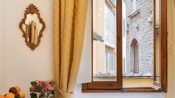A room with a wooden table, a bowl of fruit, and a vase of flowers. A window with a view of a stone building is visible. - Beautiful apartment for 4 people with A/C, WIFI and TV (Florence)