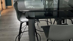 Une chambre moderne avec une table et des chaises en verre, un lit avec des rideaux bleus et une vue sur une autre pièce par une porte ouverte.