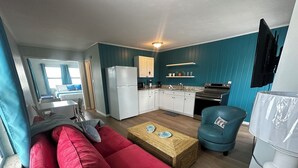 Un espace de vie compact comprenant un coin cuisine, un canapé rouge, un fauteuil bleu et une petite table à manger.