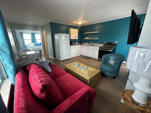 Een knus woongedeelte met een kitchenette, een rode bank, een blauwe fauteuil en een kleine eettafel.