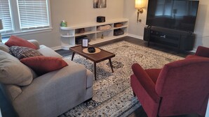 Ein Wohnzimmer mit grauem Sofa, roten Sesseln, einem Couchtisch, einem Fernseher und einer weißen Unterhaltungsanlage.