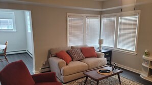 Ein Wohnzimmer mit einem beigefarbenen Sofa, einem roten Sessel und einem Couchtisch. Es gibt drei Fenster mit Jalousien, eine Lampe auf einem Beistelltisch und einen Teppich mit Muster.