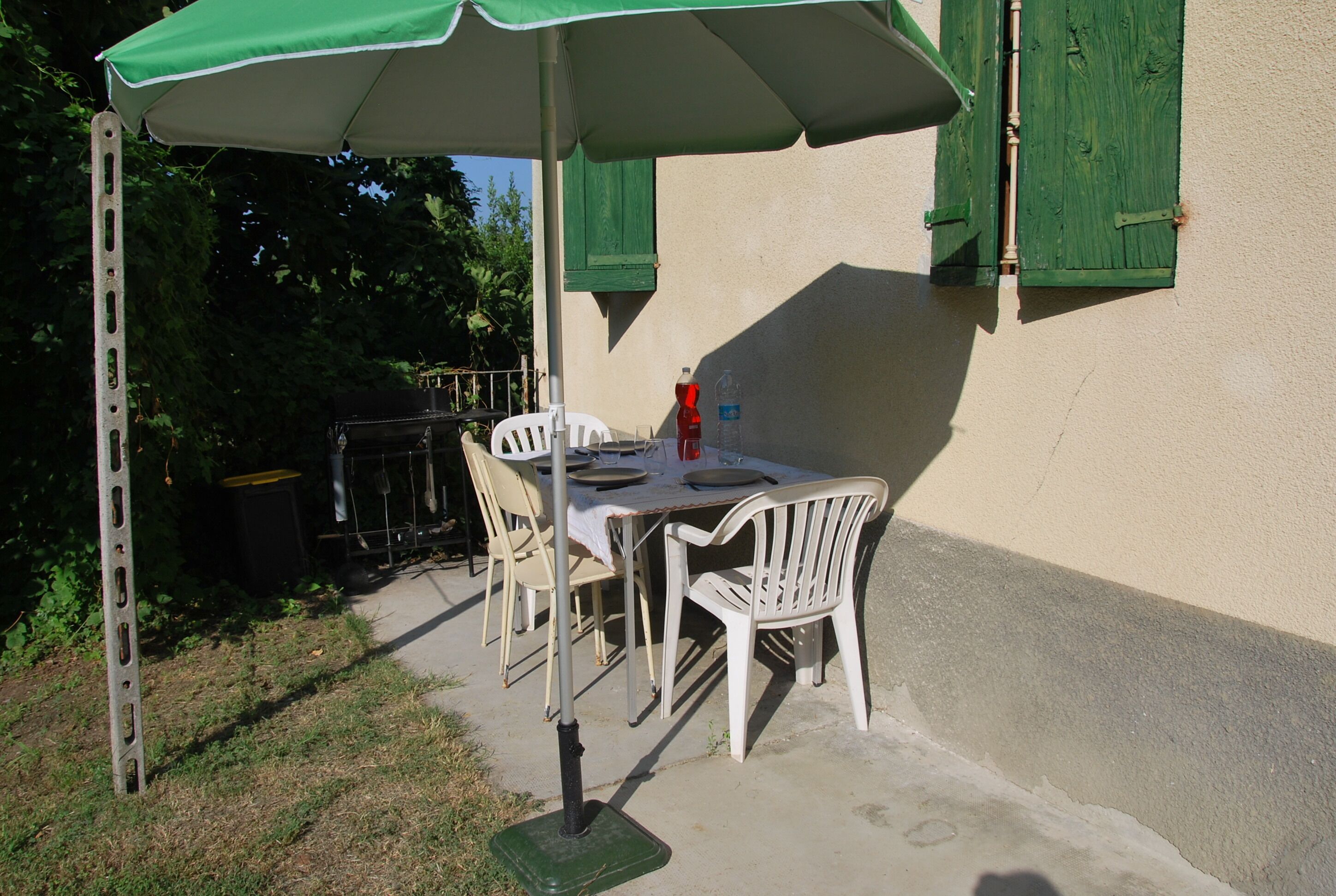 An outdoor patio with a table and chairs, a green umbrella, and a window with green shutters.