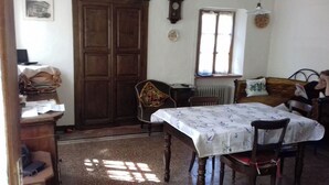Uma sala de jantar com uma mesa coberta por uma toalha de mesa florida, cadeiras de madeira, uma porta marrom, uma janela com vista e um radiador.