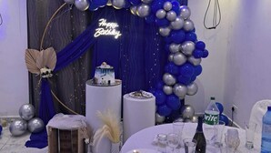 Une pièce décorée d’une arche de ballons bleus et argentés, d’un panneau néon sur lequel on peut lire « Joyeux anniversaire », et d’une table dressée pour un repas.