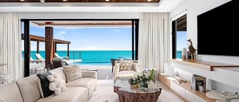Una sala de estar moderna con un ventanal grande con vista a una piscina y al mar, un sofá, una mesa de centro y un televisor montado en la pared.