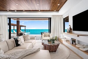 Un salon moderne doté d’une grande fenêtre donnant sur une piscine et la mer, avec un canapé, une table basse et un téléviseur fixé au mur.