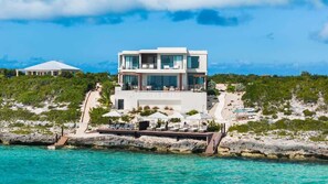 A modern two-story building with a pool area, overlooking turquoise waters and a rocky landscape.
