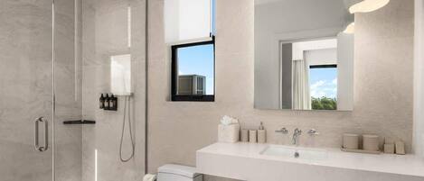 A modern bathroom with a glass-enclosed shower, a white toilet, a large mirror, and a white sink with toiletries.