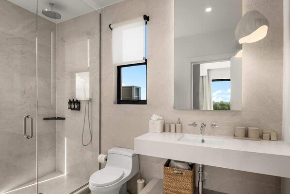 A modern bathroom with a glass-enclosed shower, a white toilet, a large mirror, and a white sink with toiletries.