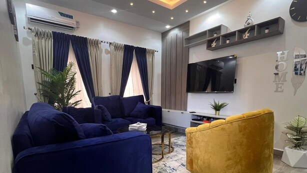A modern living room with a blue sofa set, a yellow armchair, and a flat-screen TV mounted on the wall. - Stylishly decorated 2-bedroom apartment in a serene lovely environment (Lagos)