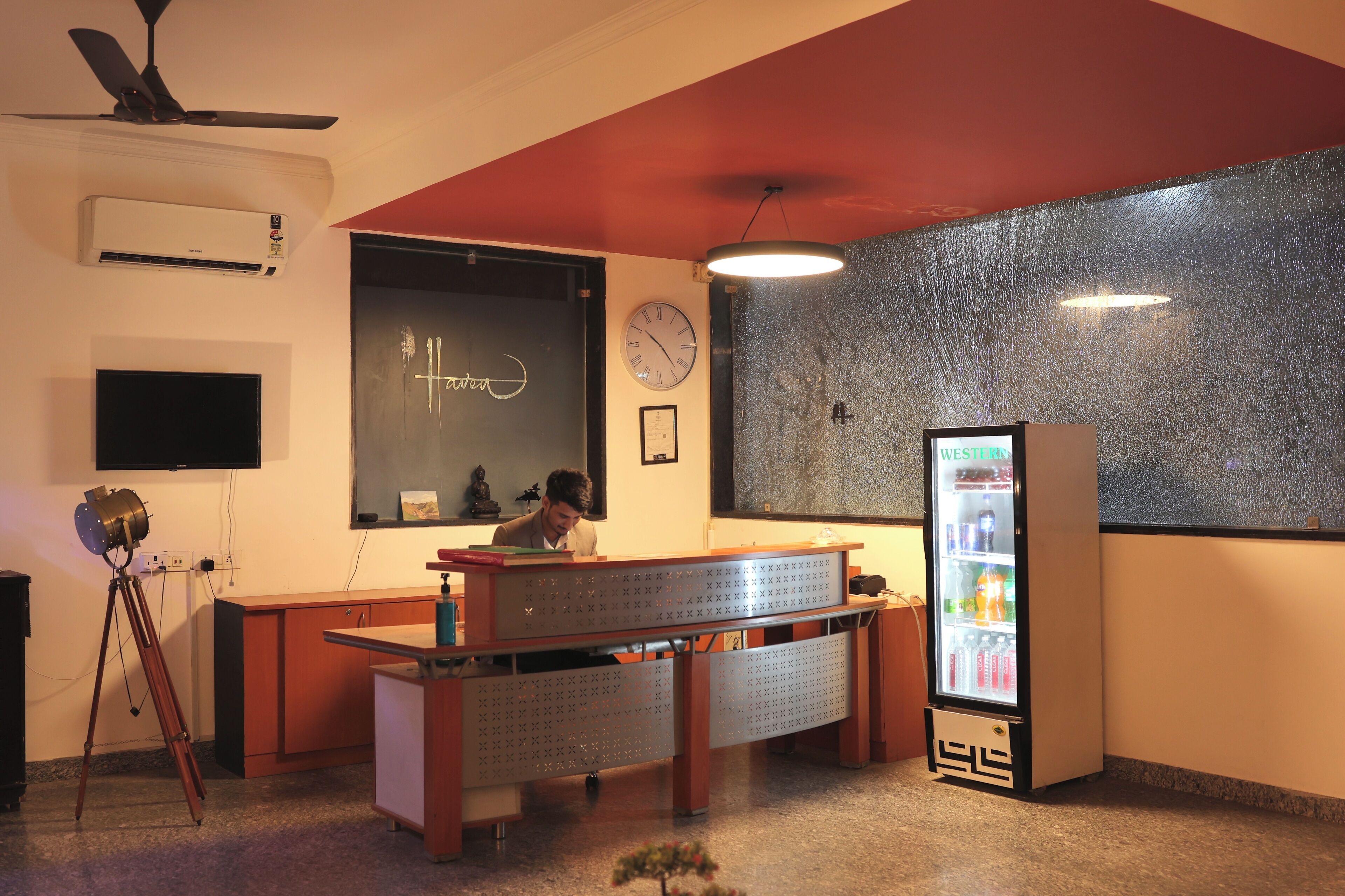 A room with a desk, a clock, a wall-mounted air conditioner, a ceiling fan, a television, and a vending machine.