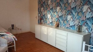 Une chambre à coucher avec un lit, une armoire blanche, une chaise en osier et un mur orné d’un motif de feuilles.