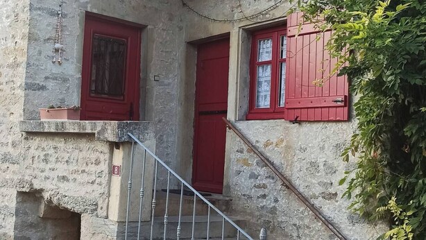 Un bâtiment en pierre avec une porte et des fenêtres rouges, un escalier en métal et un petit jardin.