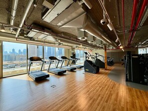 A gym with treadmills, stationary bikes, and exercise equipment, large windows with a city view, and a wooden floor.