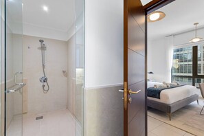 A modern hotel room with a glass-enclosed shower, a bed with a dark blue bedspread, and a large window providing a view of the city.