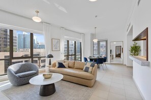 Ein modernes Wohnzimmer mit Sofa, Sessel und rundem Couchtisch. Es gibt einen Essbereich mit Tisch und Stühlen sowie große Fenster mit Blick auf die Stadt.