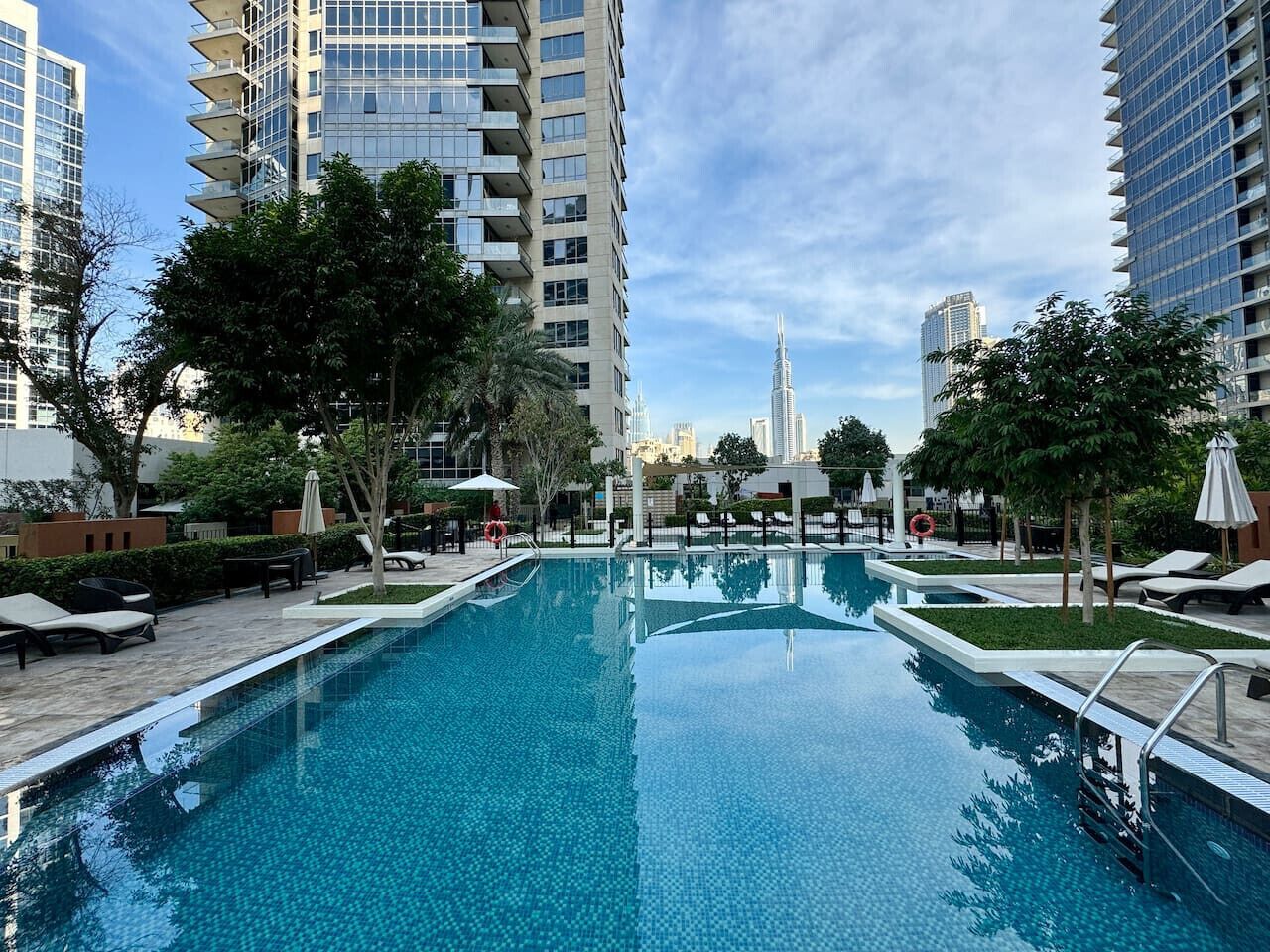 A swimming pool with lounge chairs and umbrellas, surrounded by tall buildings and trees.