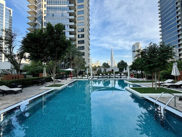 A swimming pool with lounge chairs and umbrellas, surrounded by tall buildings and trees. - Exquisite and Capacious 1BR in South Ridge 3 (Dubai)
