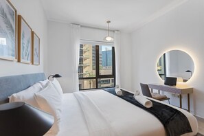 A modern hotel room with a large bed, a desk with a laptop, a round mirror, and a view of the city through the window.