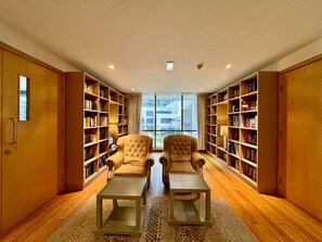 A library with two armchairs, wooden bookshelves, and a large window. - Exquisite and Capacious 1BR in South Ridge 3 (Dubai)