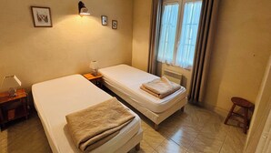 Una habitación pequeña y limpia con dos camas individuales, una mesita de noche con lámpara, una ventana con cortinas y un radiador.