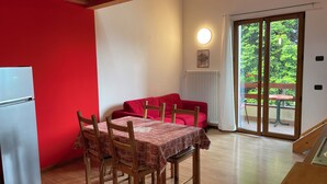 A compact room with a dining table, chairs, a red sofa, a white refrigerator, and a sliding glass door leading to an outdoor area with greenery. - App 5 Villa P. Ideal for 4 people with pool for a vacation of sports and relaxation (Malcesine)