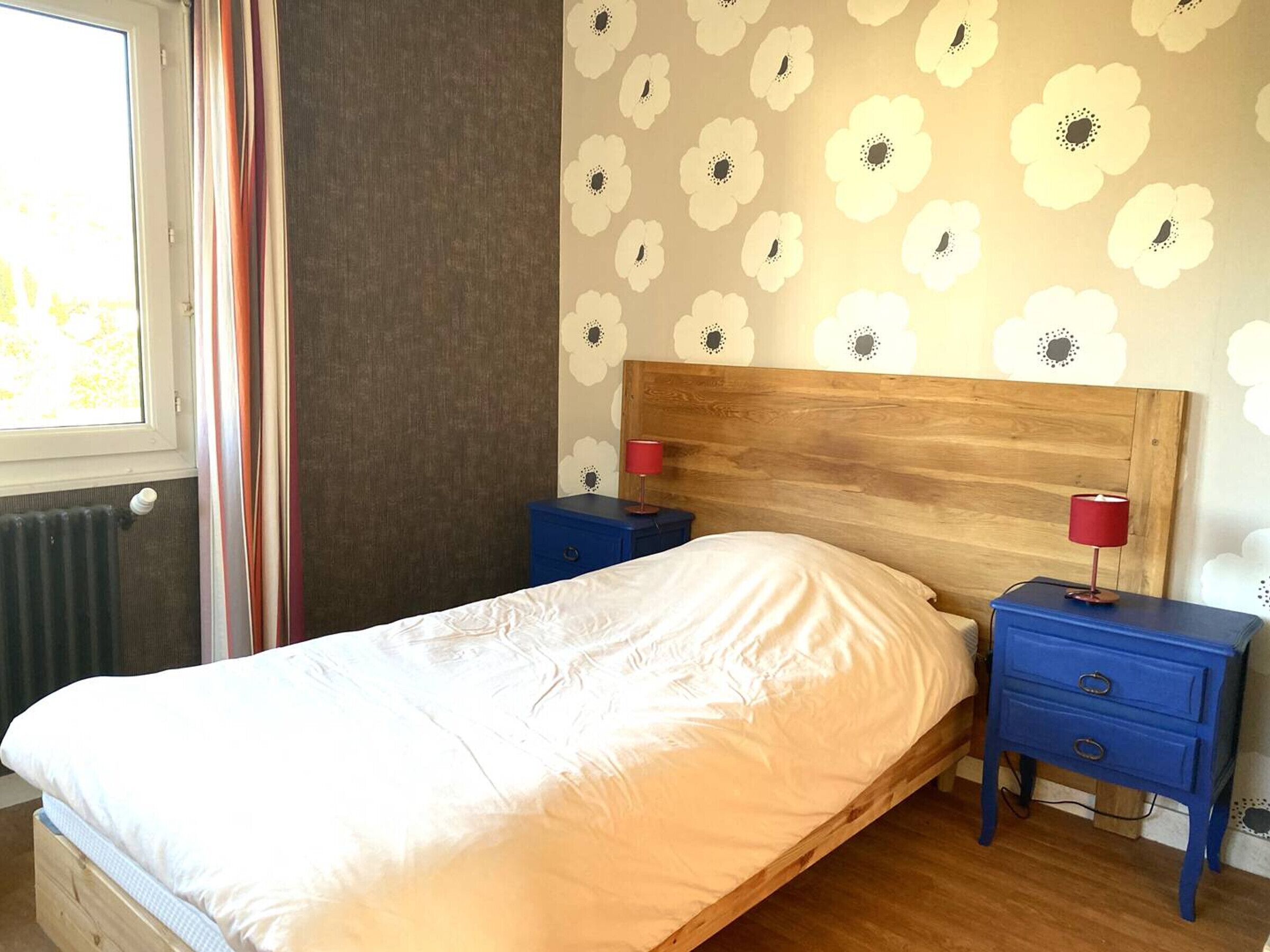 A bedroom with a wooden headboard, a bed with white bedding, two nightstands with red lamps, and a floral-patterned wall.