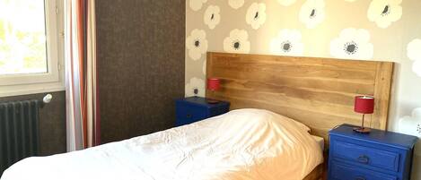 A bedroom with a wooden headboard, a bed with white bedding, two nightstands with red lamps, and a floral-patterned wall.