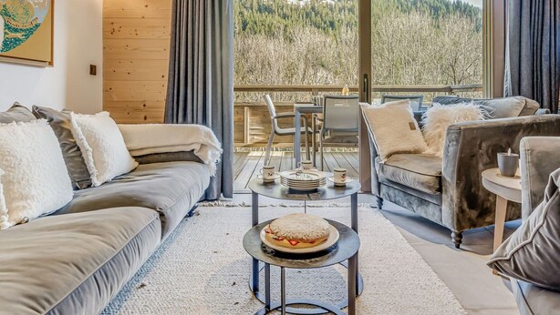 Ein gemütlicher Wohnraum mit einer Couch, einem Sessel und einem Couchtisch mit einer Schüssel Essen. Ein Balkon bietet Blick auf Bäume und Berge.
