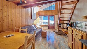 Interno di una baita di legno con zona pranzo, divano, angolo cottura con forno a microonde e vista su un paesaggio innevato attraverso ampie finestre.