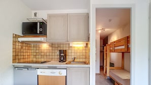 Una cocina compacta con lavavajillas, microondas y cafetera. Hay un lavabo pequeño y armarios de madera. Se ve una litera en la habitación contigua.