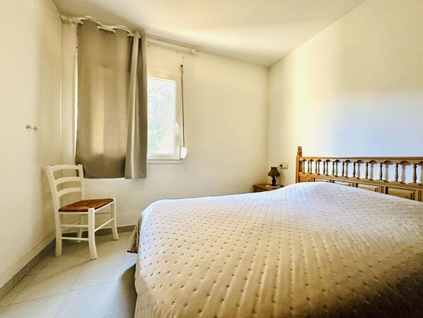 Un dormitorio con cama, una silla, una mesita de noche y un ventanal con cortinas.