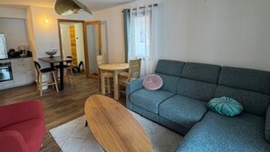 A modern living room with a grey sofa, a wooden bench, and a red chair. There is a dining area with a table and chairs, and a kitchen with a stove and cabinets.
