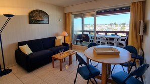 Una habitación con un sofá de color oscuro, una pequeña mesa de madera y una mesa redonda con sillas. Hay un ventanal con cortinas, una lámpara y un cuadro en la pared.