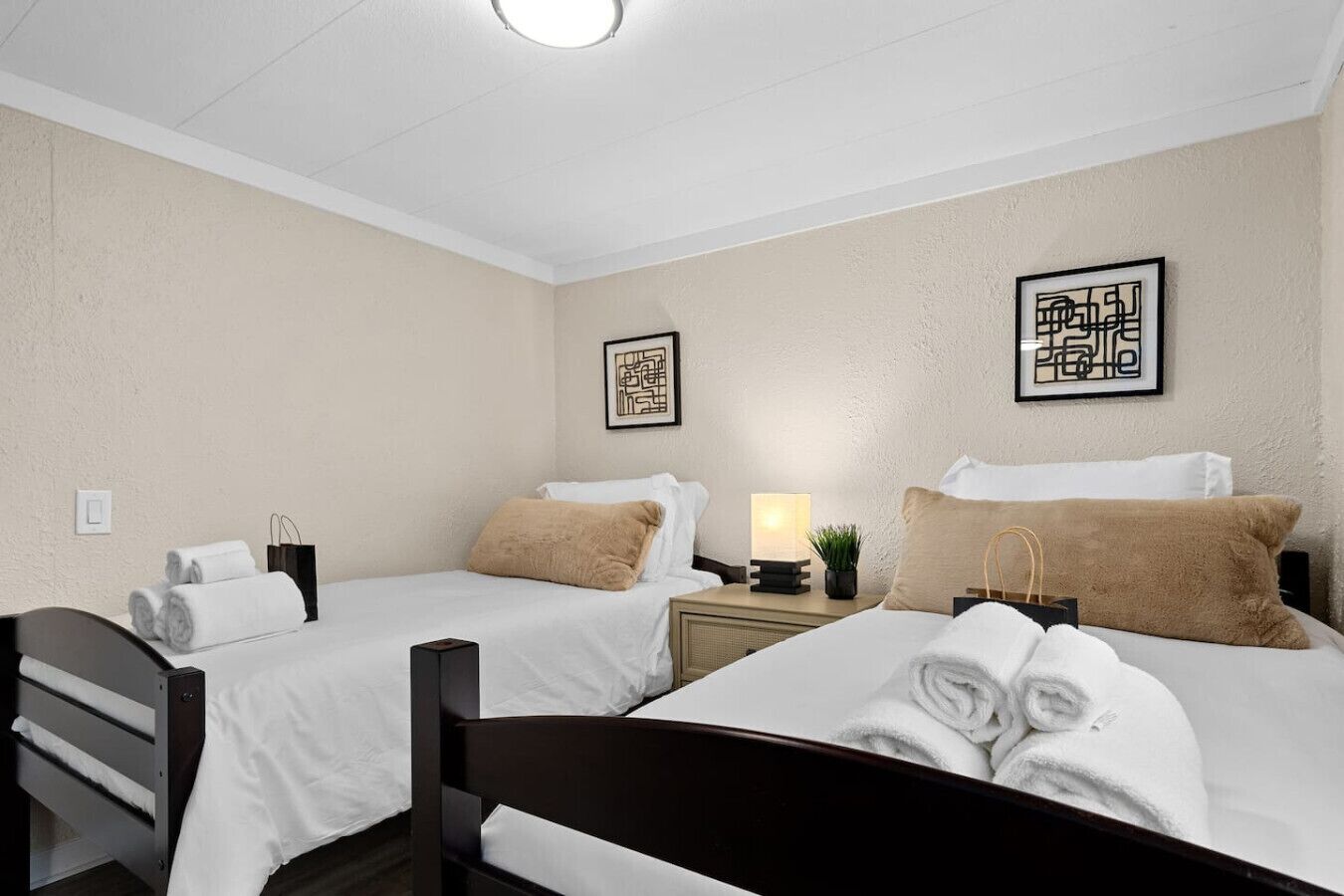 Ein Hotelzimmer mit zwei Betten, jeweils mit weißen Laken und einem braunen Zierkissen. An den Wänden befinden sich gerahmte Kunstwerke, ein Nachttisch mit einer Lampe und ein Sessel.