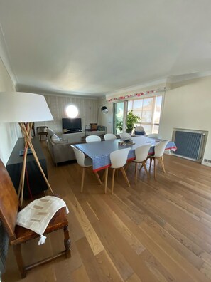 Un comedor con una mesa dispuesta para cuatro personas, rodeada de sillas blancas. Se ve una silla de madera con mantel blanco en primer plano.