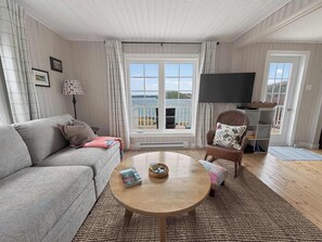 A living room with a sofa, a coffee table, a wicker chair, a television, and a view of the sea through a large window. - Seaside Serendipity Waterfront Cottage near Twillingate (Merritt's Harbour)