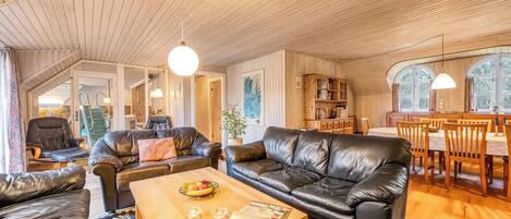 A spacious living room with a wooden ceiling, leather sofas, a wooden coffee table, and a dining area with a wooden table and chairs.