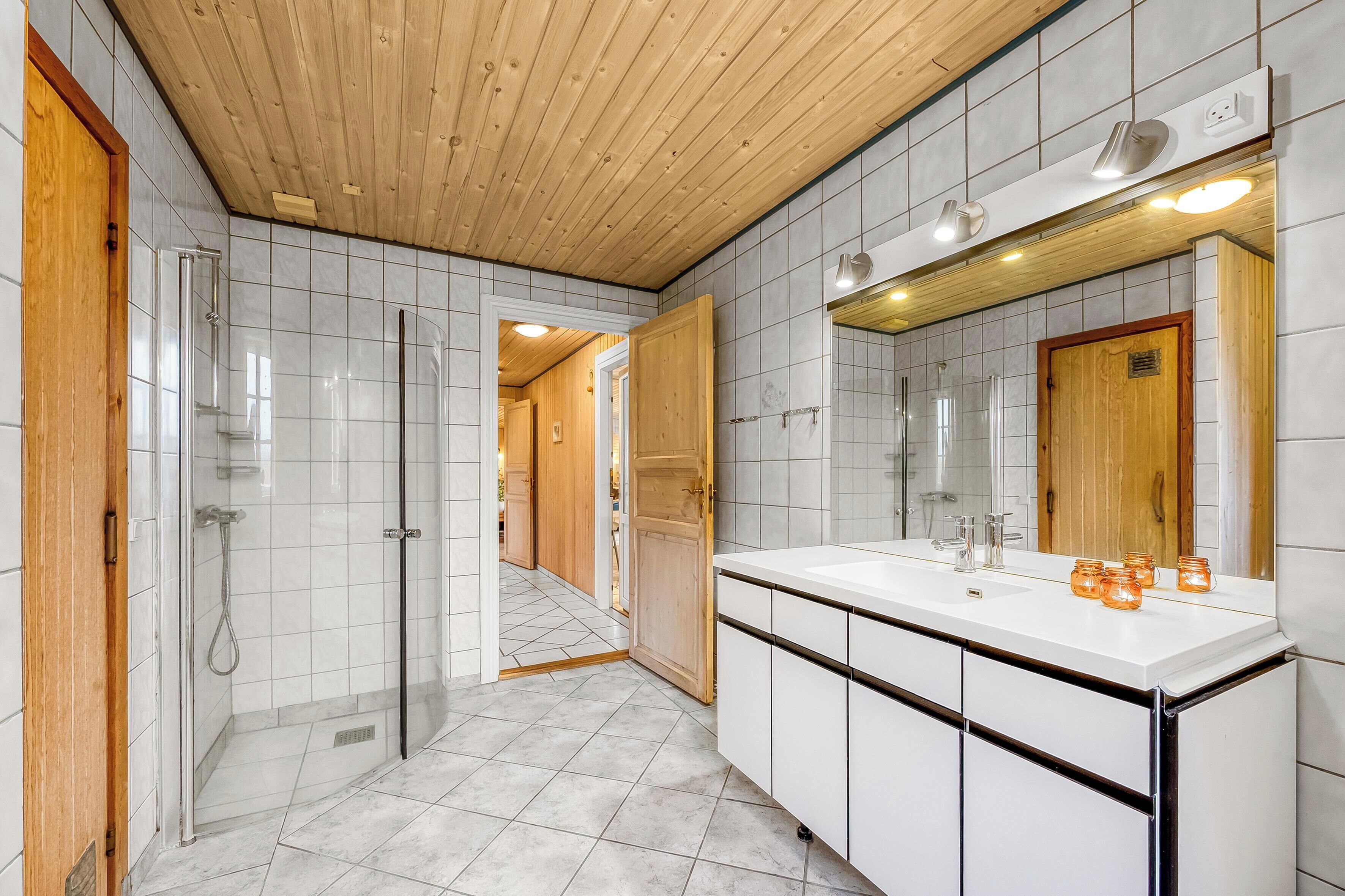 Une salle de bain moderne avec un plafond en bois, des carreaux blancs, une cabine de douche en verre et un grand miroir au-dessus du lavabo.