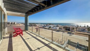 A balcony with a view of the beach and ocean, red chairs, and a clear sky.