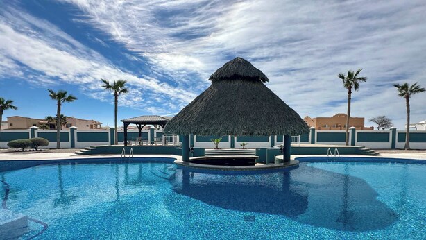 A swimming pool with a thatched-roof gazebo, palm trees, and a clear sky.