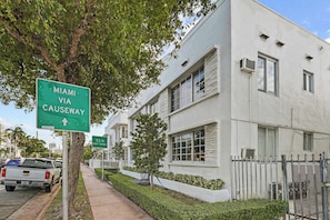 A residential building with a white exterior, a tree-lined street, and a green directional sign indicating 'Miami Via Causeway'.