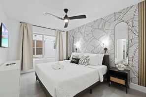 Ein Schlafzimmer mit Bett, Deckenventilator, Nachttisch, Spiegel und einem Fenster mit Vorhängen.