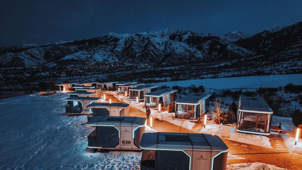 En række moderne hytter med sne på taget og oplyste interiører, foran et bjergrigt landskab.