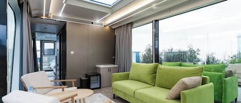 A modern interior with a green sofa, a wooden chair, a small table, and a large window offering a view of the outdoors.