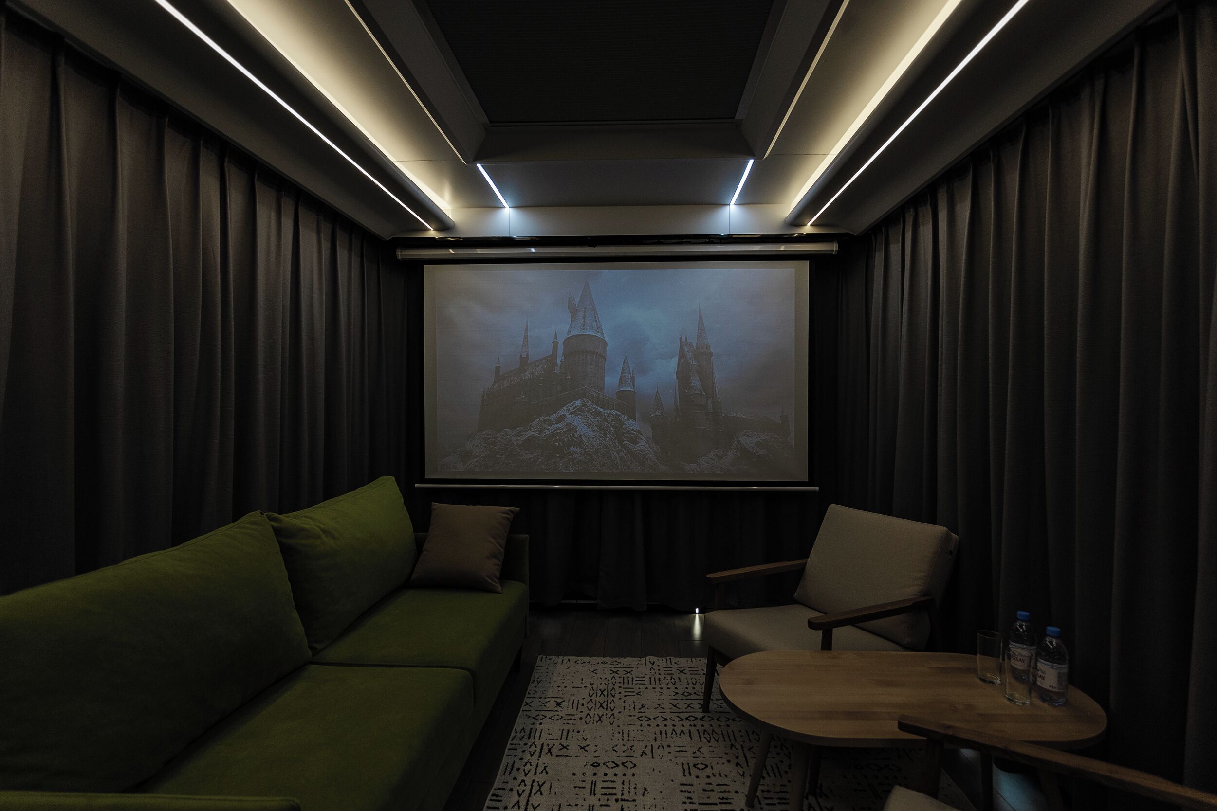 Ein dunkles Zimmer mit großem Bildschirm, grünem Sofa und einem kleinen Tisch mit zwei Flaschen.