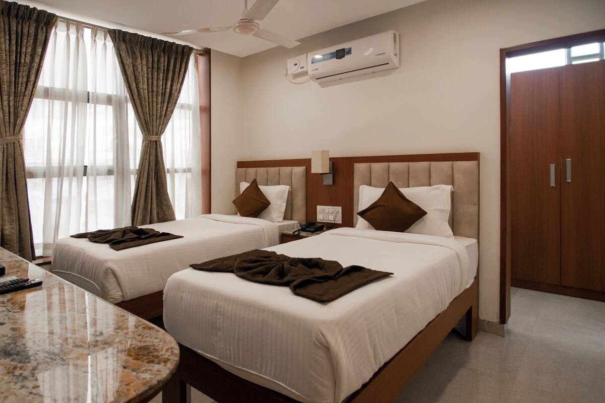 Ein Hotelzimmer mit zwei Betten, einem Marmortisch, einem Deckenventilator, einer Klimaanlage und einem Holzschrank.